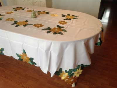 A dining table features the XMas Tablecloth Gold Poinsettia Emerald Green Leaf Appliqué Oblong, showcasing intricate yellow and green floral patterns. In the center, a small decorative tree is displayed. The table rests on a hardwood floor.