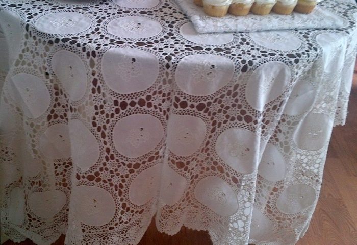 A round table is draped with the Wedding Ring WhiteWork Rose Tablecloth – Handcrafted Heirloom Lace, its intricate circular patterns displayed beneath a tray of dessert cupcakes. A wooden floor enhances the elegant, warm setting.