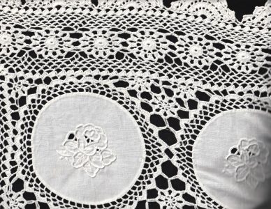 A close-up of the Wedding Ring WhiteWork Rose Tablecloth, a handcrafted heirloom lace with intricate floral patterns and embroidered white circles, beautifully displayed on a black background—perfect for elegant wedding décor.