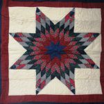 Introducing the Tartan Lone Star Quilted Tablecloth & Wall Hanging, featuring an intricate eight-pointed star design crafted with plaid fabric in shades of red, gray, and black on a cream background. The star is elegantly framed with maroon and dark blue fabric strips.
