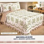 A bedroom features a bed adorned with an Irish Green & White Double Wedding Ring Handmade Quilt, showcasing a beautiful floral pattern. The set is complete with matching pillow shams. A wooden nightstand with a lamp and a chair are positioned next to the bed. Text reads "Heirloom 100% Hand Quilted.