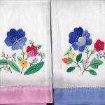 Two white hand towels with embroidered colorful flowers and green leaves. The left towel has a pink edge, and the right towel has a blue edge. Both feature similar floral patterns in purple, pink, yellow, blue, and red.