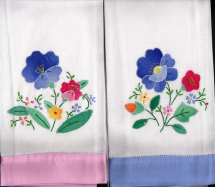 Two white hand towels with embroidered colorful flowers and green leaves. The left towel has a pink edge, and the right towel has a blue edge. Both feature similar floral patterns in purple, pink, yellow, blue, and red.