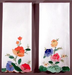 Set of two white guest towels with appliqué hand-stitched Floral or Wild Rose designs. Each features unique arrangements of red, blue, yellow, and pink flowers with green leaves on a plain background.
