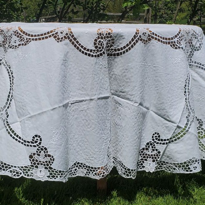 The Daffodils Reticella Lace Round Tablecloth, featuring linen needle embroidery, is displayed outdoors on grass with trees and a fence in the background.
