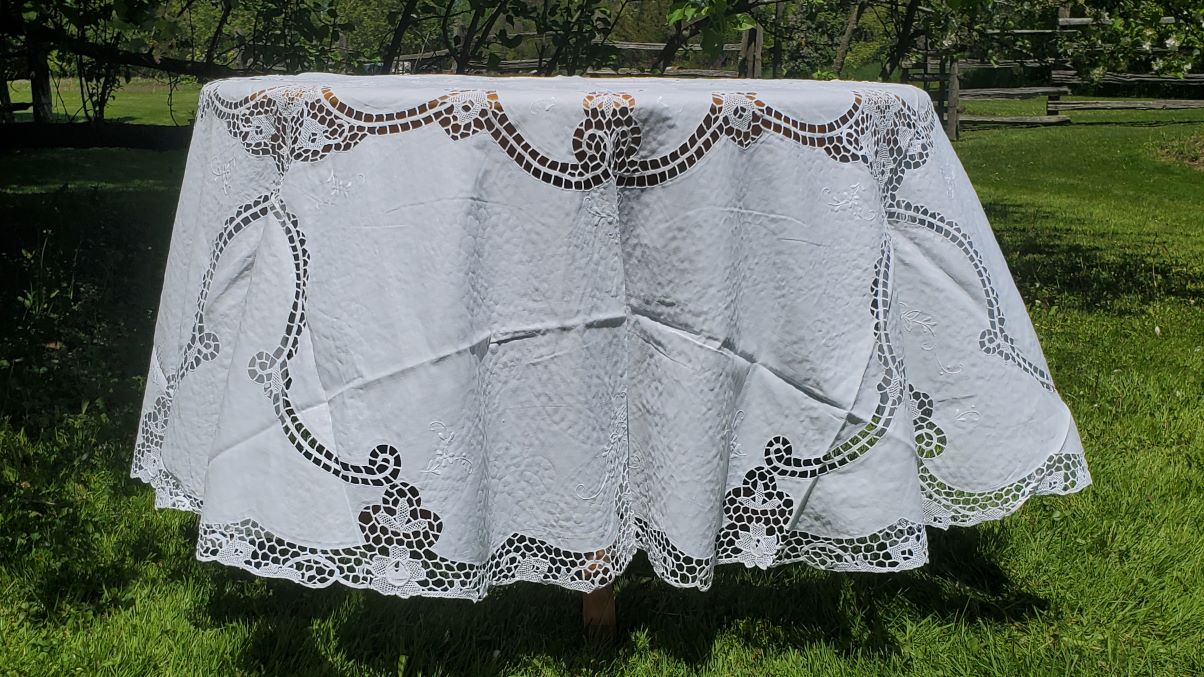 The Daffodils Reticella Lace Round Tablecloth, featuring linen needle embroidery, is displayed outdoors on grass with trees and a fence in the background.