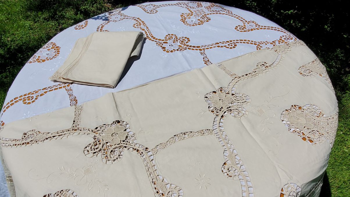 Outdoors on green grass, a table is draped with the Daffodils Reticella Lace Round Tablecloth Linen Needle Embroidery. On top are a folded beige cloth and a matching lace table runner.