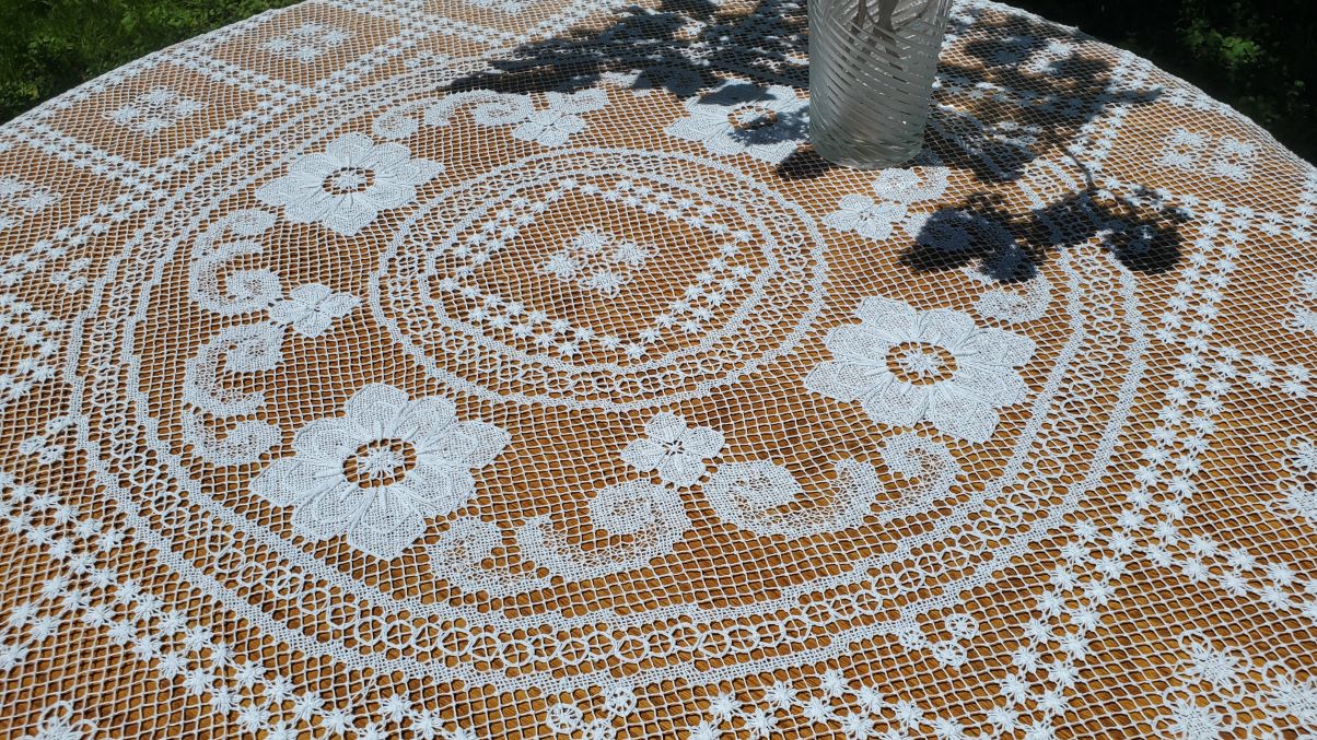 A close-up of the Fine Table Linens: Tuscany Lace Hand Knotted Italian Modano Round tablecloth, showcasing its detailed floral and geometric lace. A glass vase casts a shadow on the sunlit, grassy background.