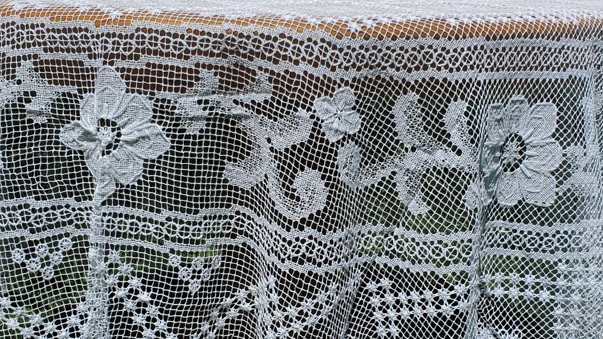 The Fine Table Linens: Tuscany Lace Hand Knotted Italian Modano Round tablecloth features intricate floral and geometric patterns on delicate mesh, allowing the surface beneath to show through.