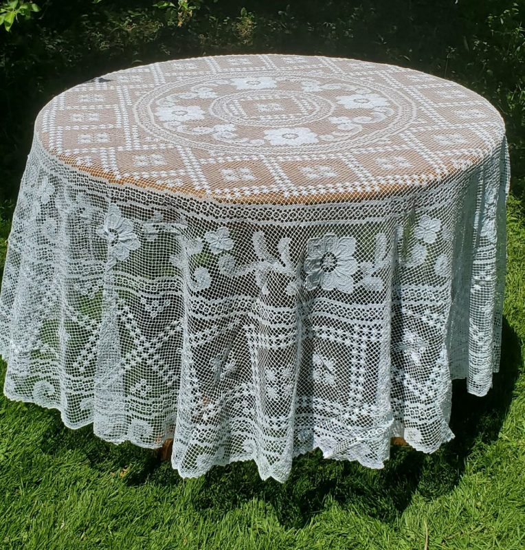 A Fine Table Linens: Tuscany Lace Hand Knotted Italian Modano Round tablecloth covers a round table set on green grass outdoors, sunlight casting shadows and greenery in the background.