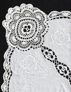 A close-up of the Crochet Lace Roses 4 Corners Tea Tablecloth in white, showcasing embroidered flowers and intricate crocheted edges, displayed on a black background.