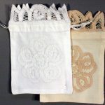 A pair of fabric pouches designed with Battenburg lace, one in white and the other in beige, each featuring intricate lace patterns on the front. Both come with drawstrings and a decorative lace trim at the top.