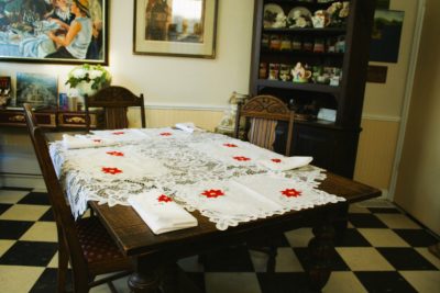 A dining room with a checkered floor features a wooden table dressed with Christmas Poinsettia & Battenburg Lace Cotton Tray Mats, white napkins with red stars, wooden chairs, shelves, and festive art, creating a cozy holiday scene.