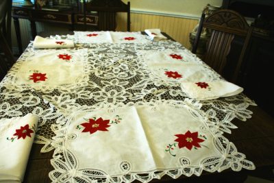 A dining table set features two Christmas Poinsettia & Battenburg Lace Cotton Tray Mats, each adorned with ornate lace and red poinsettia designs. Folded napkins are neatly placed at each seat.