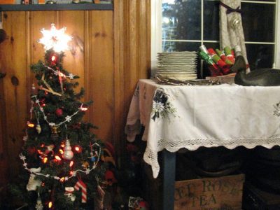 A small, decorated Christmas tree with lights stands beside a table covered with a Moonlight Christmas Blue & White Candle Crochet Lace Trim Oblong tablecloth. The table holds a stack of plates and festive Christmas crackers, while a wooden crate labeled "Rose Tea" is visible below.