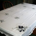 The dining table is adorned with the Moonlight Christmas Blue & White Candle Crochet Lace Trim Oblong tablecloth, showcasing intricate embroidered floral patterns and festive designs. Small bouquets tied with midnight blue ribbons embellish each corner and the center. Wooden chairs surround the table.