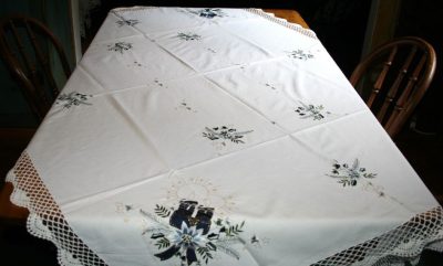 A rectangular dining table is adorned with the Moonlight Christmas Blue & White Candle Crochet Lace Trim Oblong tablecloth, which showcases embroidered floral patterns and a candle motif. The lace trim enhances its elegance, and two wooden chairs are positioned at opposite ends of the table.