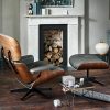 A stylish living room features a mid-century modern lounge chair with matching ottoman, both with wooden frames and black cushions. A small table holds wrapped gifts, while a white marble fireplace with stacked logs and various décor items enhances the cozy ambiance.