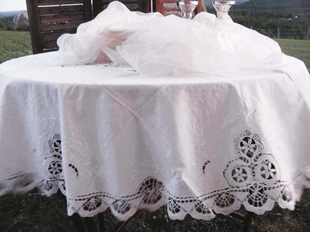 Outdoors on grass with hills and trees behind, a round table is draped in a Cluny Lace Bobbin Lace Round Tablecloth Handmade Lace Edging White. Two silver cups rest on it near wooden panels.