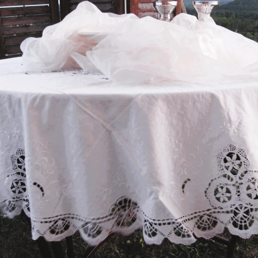 Outdoors on grass with hills and trees behind, a round table is draped in a Cluny Lace Bobbin Lace Round Tablecloth Handmade Lace Edging White. Two silver cups rest on it near wooden panels.