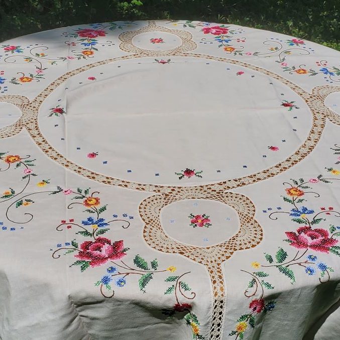 The Fine Table Linen: Royal Albert "Old Country Rose" CrossStitch Round features colorful floral embroidery and lace accents, displayed outdoors on grass with sunlight highlighting its elegant white fabric.