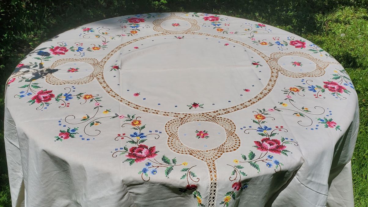 The Fine Table Linen: Royal Albert "Old Country Rose" CrossStitch Round features colorful floral embroidery and lace accents, displayed outdoors on grass with sunlight highlighting its elegant white fabric.