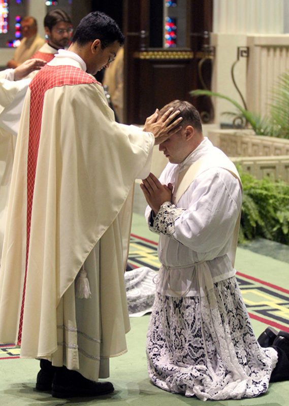During a religious ceremony inside a church, a priest in white robes—Tradition Lithurgical ALB: DIY-Solid Battenburg Lace Oblong Tablecloth—places his hands on a praying man in ornate vestments.