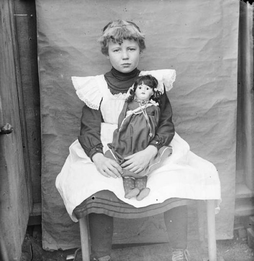 A black and white photograph shows a young girl with short hair dressed in a dark blouse with a white apron. She is seated on a wooden chair, holding a doll in her lap. The background is a plain, textured curtain. The girl has a somber expression.