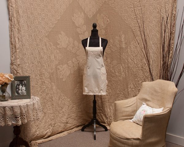 A black mannequin dressed in a beige apron stands in front of an ornate beige tapestry. A vintage armchair with a floral pillow is on the right. On the left, a small round table with a lace cloth has a vase with flowers and a framed photo. Dried branches are in the background.