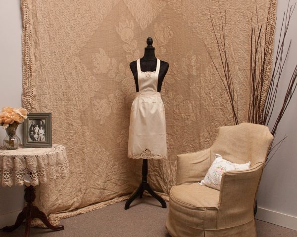 A vintage room setup with a mannequin dressed in a white apron, a beige chair with a floral pillow, and a small table covered with a lace cloth. The table holds a framed black-and-white photo and a vase of flowers. The backdrop and decor feature intricate lace patterns.