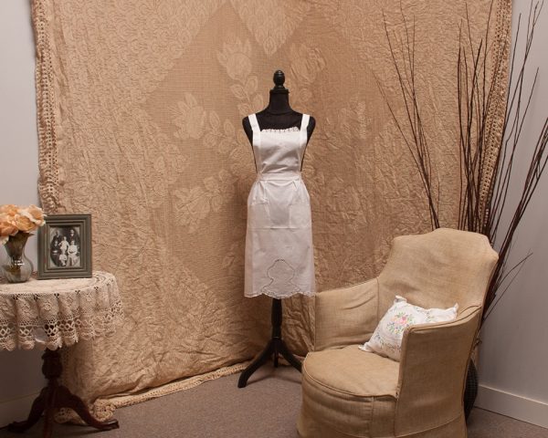 A vintage dress form draped with an elegant white apron stands in the corner of a room. Behind it, a beige, intricately patterned backdrop hangs. A beige armchair with a white floral cushion and a lace-covered table holding a framed black-and-white photo complete the scene.