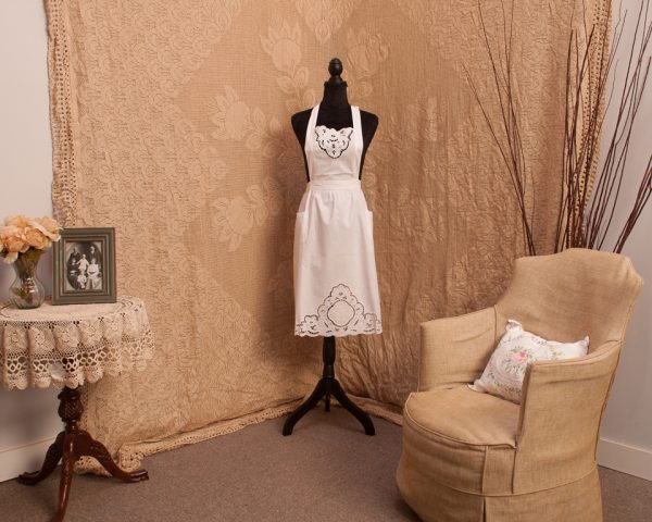 A vintage-style room features a mannequin dressed in a white apron in the center. To the left, a lace-covered table displays a framed photo and floral arrangement. A beige armchair with an embroidered pillow sits to the right, against a backdrop of patterned fabric.