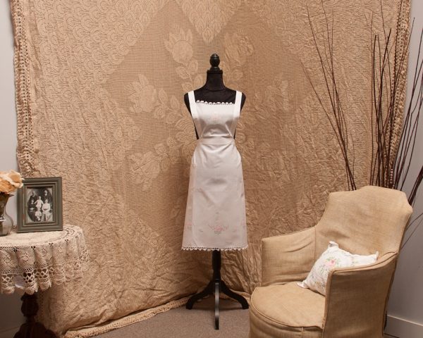 A vintage-style room featuring a white embroidered apron on a black mannequin. The background has a beige, floral-patterned tapestry. A beige armchair with a floral cushion, a small round table with lace cloth, framed photo, and a vase with flowers complete the scene.