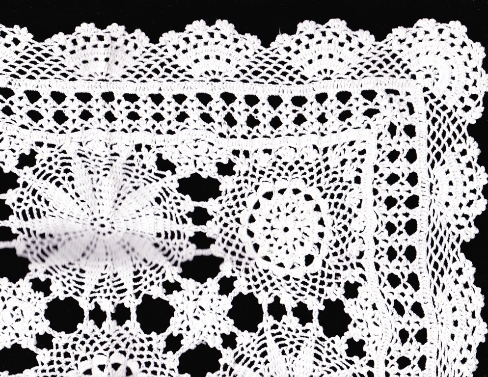 Close-up of a detailed white crocheted lace fabric with intricate floral patterns on a black background. The lace features a mix of starburst, circular, and scalloped edge designs.