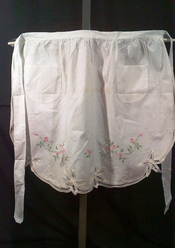 A white apron with decorative embroidery. The apron features two pockets and a floral design with pink roses and green leaves near the bottom hem. The hem is scalloped and adorned with lace trim. The apron is tied at the waist with long fabric straps.