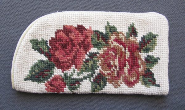 A small, rectangular, white fabric bag with a cross-stitched design of red and pink roses, surrounded by green leaves, on one side. The bag sits on a dark gray textured surface.