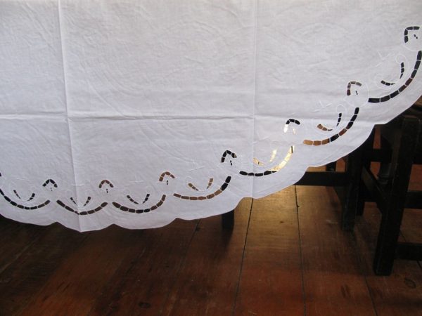 A close-up of a white tablecloth with an intricate cut-out tulips pattern along the scalloped edge. The tablecloth is draped over a wooden table, and the floor is made of polished wood planks.