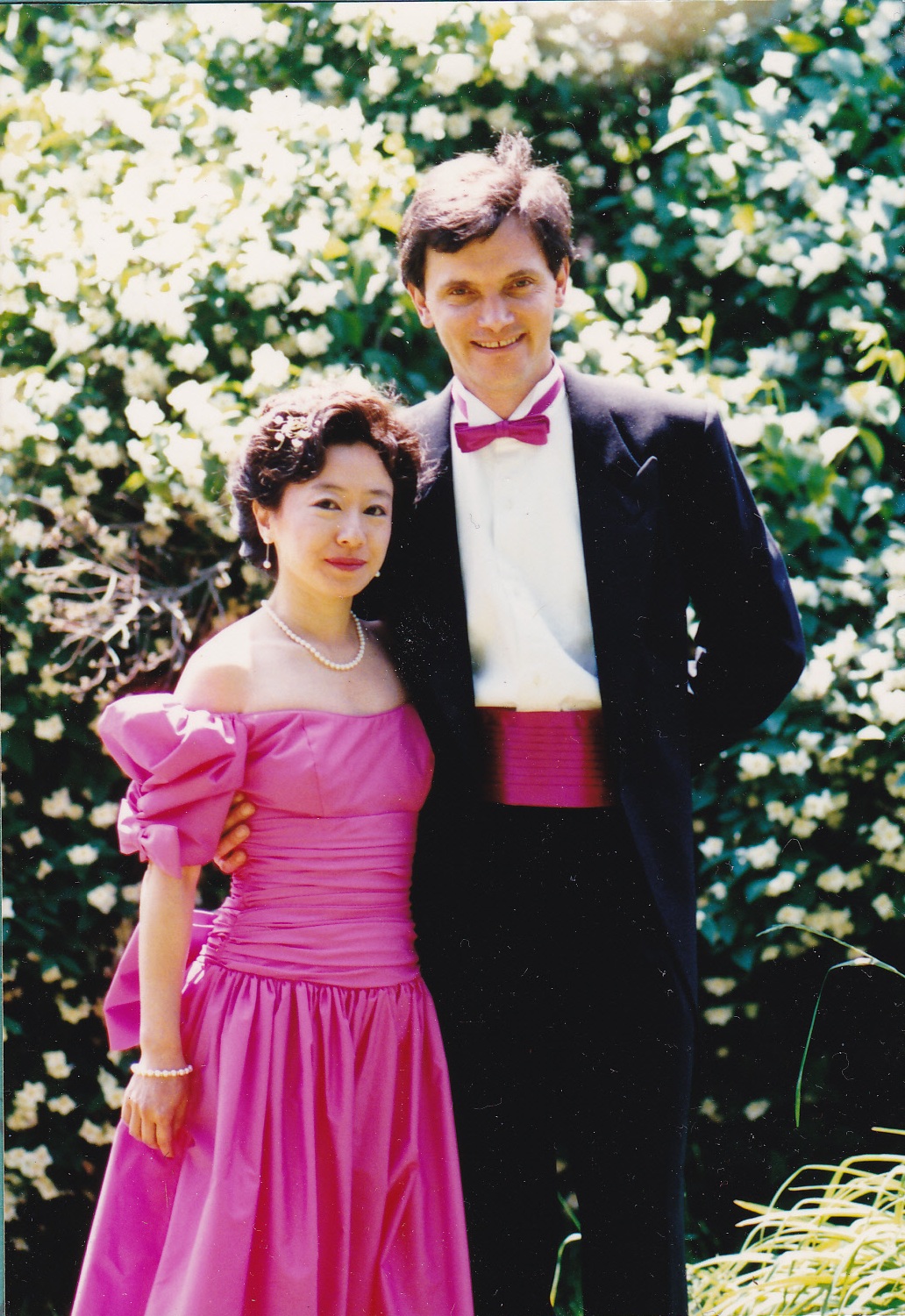 A woman in a pink off-shoulder dress with puff sleeves and a matching belt, standing with a man in a black tuxedo with a pink cummerbund and bow tie. They are outdoors with greenery in the background, smiling and posing for the photo.