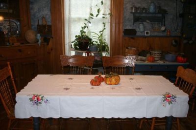 A cozy dining room with a wooden table covered by a floral tablecloth. On the table are decorative pumpkins. The room features wooden chairs, a sideboard with dishes, plants, and rustic decor.