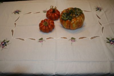 Three ceramic pumpkins are displayed on a white embroidered tablecloth with floral designs, set on a table.