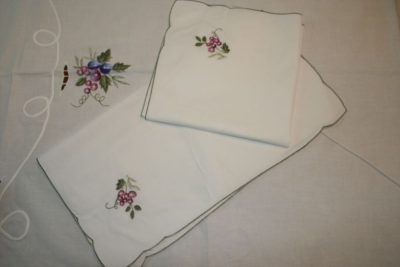 A set of white linen napkins with embroidered purple flowers and green leaves is neatly folded and laid on a matching tablecloth. The embroidery adds a delicate touch to the otherwise plain fabric.