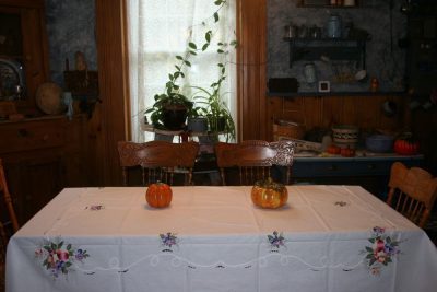 A cozy kitchen scene with a wooden table covered by an embroidered white tablecloth. Three small pumpkins serve as decoration. In the background are wooden cabinets, shelves with various kitchen items, and a plant by the window filtering soft light.