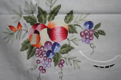 A vibrant embroidered design on fabric featuring colorful fruits like apples and grapes, surrounded by green leaves. A small red butterfly is also included in the intricate stitching.