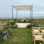 An outdoor wedding setup featuring the unique Wedding Chuppach Canopy, a SnowFlake Crochet Bed Cover overlooks the ocean, with greenery and white flowers enhancing the ambiance. Wooden chairs are meticulously placed on a lush grassy lawn, while a few boats can be seen in the distance beneath a cloudy sky.