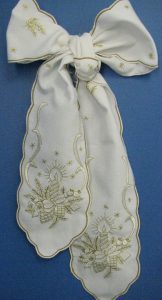 A decorative white bow with intricate gold embroidery, featuring detailed designs of candles and stars. The bow is displayed against a plain blue background.