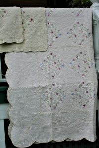 The Fairytale Flower Garden Quilt Cotton Embroidery Whole Cloth, adorned with diamond-shaped embroidered flowers and scalloped edges, elegantly hangs on a railing. A matching pillow is draped beside it.