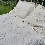 A white metal bed frame stands on grass, adorned with the Fairytale Flower Garden Quilt Cotton Embroidery Whole Cloth and two pillows. The quilt and pillows are intricately embroidered with delicate floral designs in light pink, blue, and green hues. Verdant greenery is visible in the background, suggesting an outdoor setting.