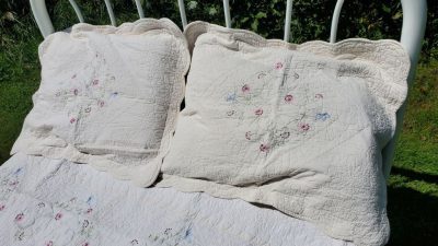 A pair of cream-colored quilted pillows and a matching Fairytale Flower Garden Quilt, adorned with floral embroidery, are displayed on an outdoor metal bed frame. Sunlight casts shadows, accentuating the detailed patterns of the quilt against a backdrop of greenery.