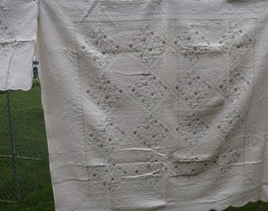 The Fairytale Flower Garden Quilt, featuring an exquisite floral diamond pattern, is hanging on a clothesline. Its intricate stitching and delicate pink flower design stand out against the backdrop of green grass and a chain-link fence.