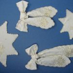 A set of white and gold-embroidered ornaments on a blue background, including two star-shaped pieces and two bow-shaped decorations. The embroidery features floral patterns and delicate details.